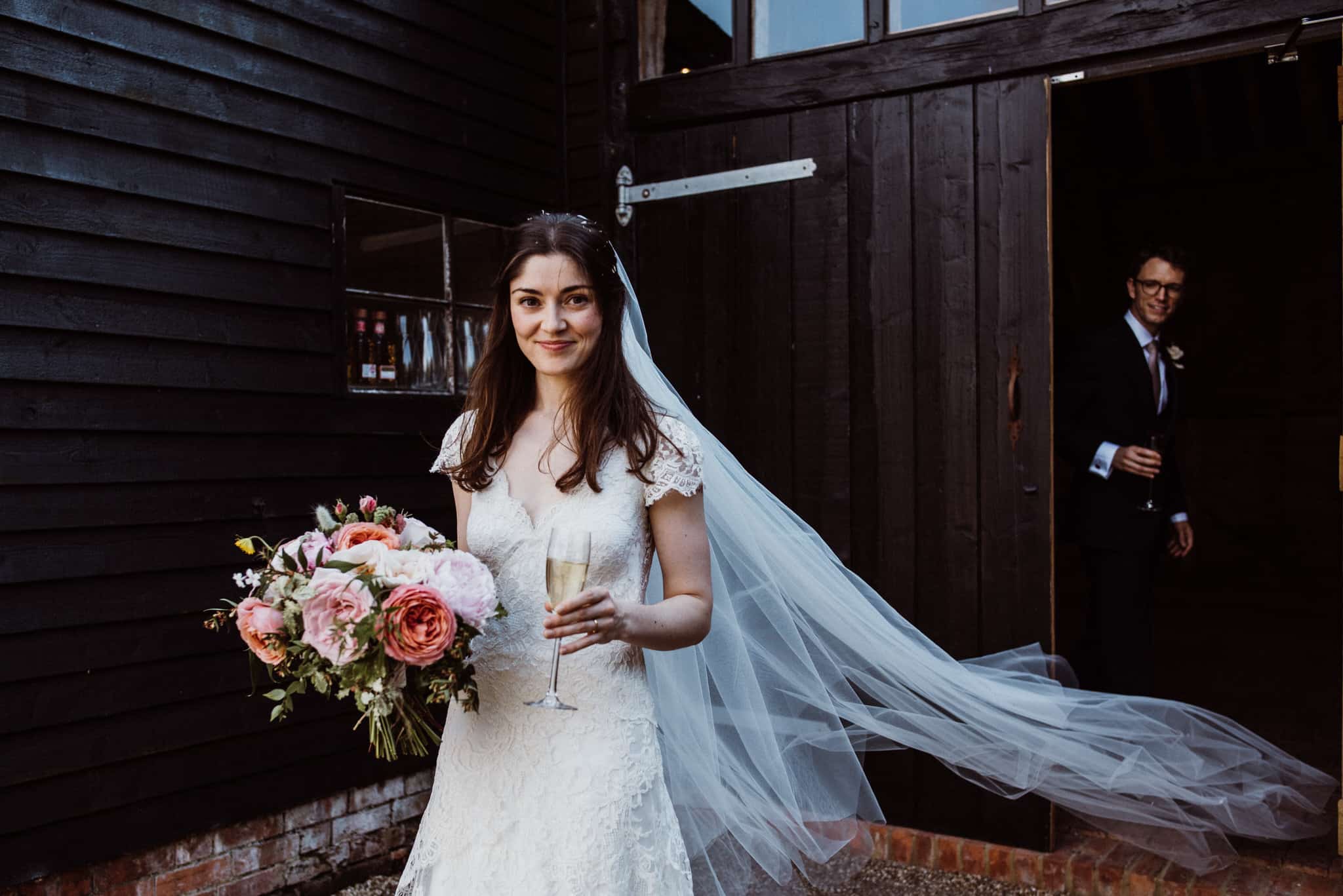 Bride holding bouquet and champagne outside High Billinghurst Farm barn wedding venue in Surrey
